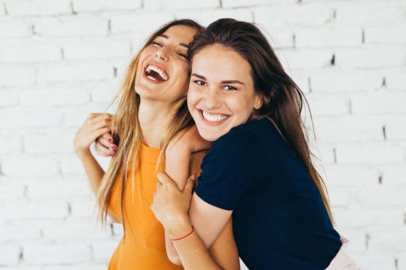 Two young women laughing and hugging against a white brick wall Advanced Orthodontics in Kent, WA
